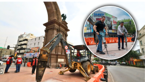 Construction work around a historic monument with an excavator, orange barriers, and workers in red vests overseeing the site.