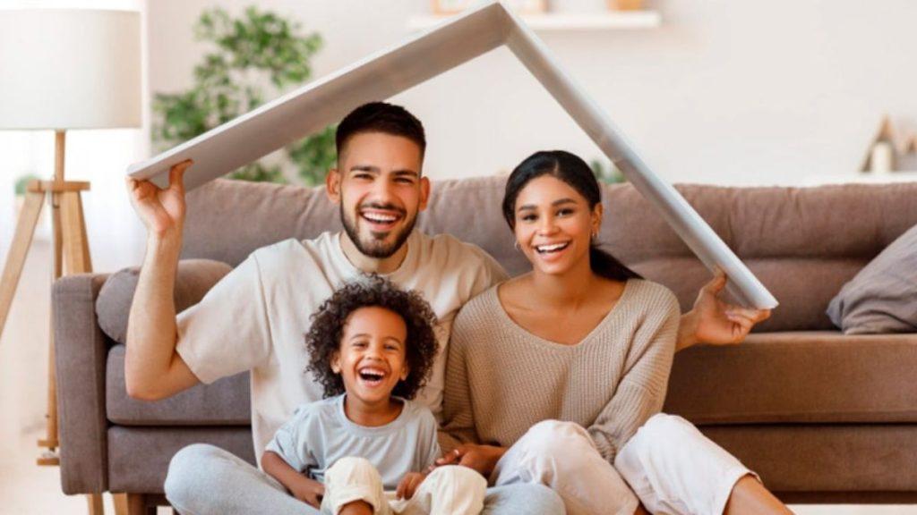 a Happy Family of Three Sits on the Living Room Floor Under a Makeshift Cardboard Roof, Smiling at the Camera. - Apoyo Regio