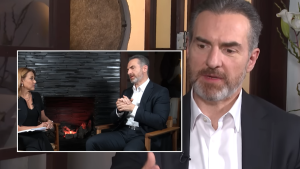 Interview scene: man in a dark suit speaks while a female host sits beside him, with a stone fireplace background in the inset frame.