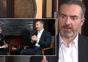 Interview scene: man in a dark suit speaks while a female host sits beside him, with a stone fireplace background in the inset frame.