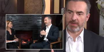 Interview Scene: Man in a Dark Suit Speaks While a Female Host Sits Beside Him, with a Stone Fireplace Background in the Inset Frame. - Apoyo Regio