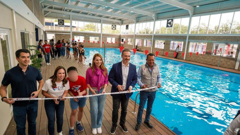 Group of Six Adults and a Child Cut a White Ribbon at a Poolside Ribbon-cutting Ceremony Indoors, with Red Pennant Banners Overhead and Spectators in the Background. - Apoyo Regio
