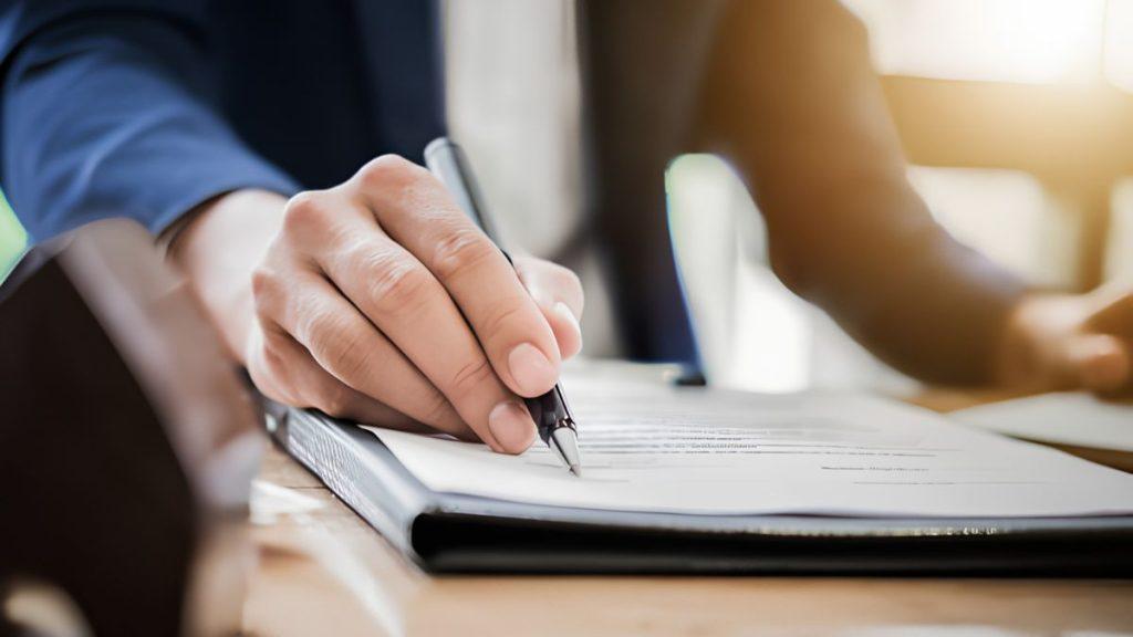 Person in a Suit Signs a Document with a Pen on a Clipboard at a Desk. - Apoyo Regio