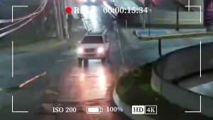 Dashcam view of a white SUV driving through a wet, rain-soaked street at night, headlights on, reflections on the flooded road.