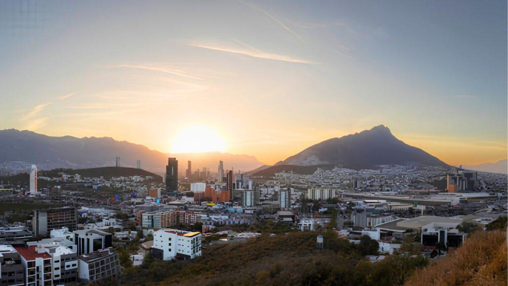 Quantium-desarrollos-monterrey-ciudad-cerros-desarrollo-urbano - Apoyo Regio Sunset over a Dense Cityscape with Tall Buildings and a Large Mountain in the Background in the Distance, Horizon Glowing Orange for Dusk. - Apoyo Regio