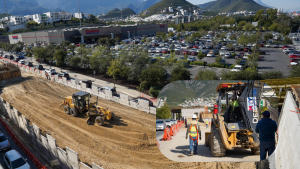 Construction site excavation with a large dirt ramp next to a Costco parking lot; heavy machinery at work and a fenced area nearby.