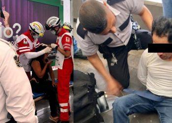Two emergency responders in red and white uniforms treat a seated person on a stretcher; a security officer watches nearby in a public setting.