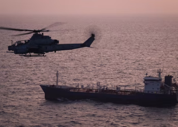 Military helicopter hovering above a tanker ship on calm sea at sunset, pinkish sky reflecting on water