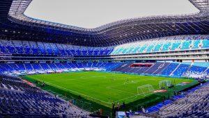 Interior of a modern soccer stadium with a green pitch and blue seating surrounding an empty arena under a curved roof.