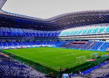 Interior of a modern soccer stadium with a green pitch and blue seating surrounding an empty arena under a curved roof.