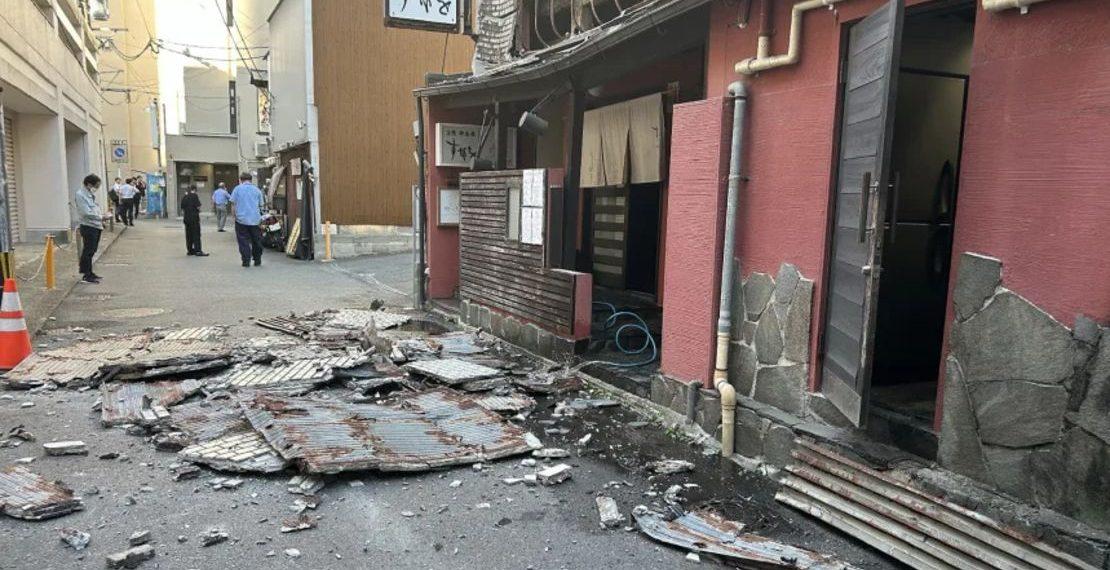 Debris and Collapsed Roofing Material Cover a Narrow Street Beside a Damaged Red Building, with a Few Bystanders in the Distance. - Apoyo Regio