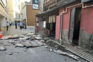 Debris and collapsed roofing material cover a narrow street beside a damaged red building, with a few bystanders in the distance.
