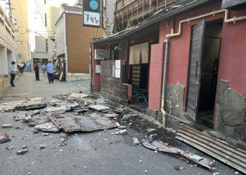 Debris and collapsed roofing material cover a narrow street beside a damaged red building, with a few bystanders in the distance.