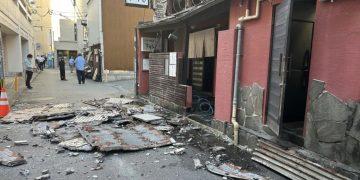 Debris and Collapsed Roofing Material Cover a Narrow Street Beside a Damaged Red Building, with a Few Bystanders in the Distance. - Apoyo Regio