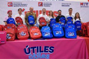 Group of eight people posing with red and blue MTY backpacks for a school supplies drive, standing behind a pink 'ÚTILES' banner.