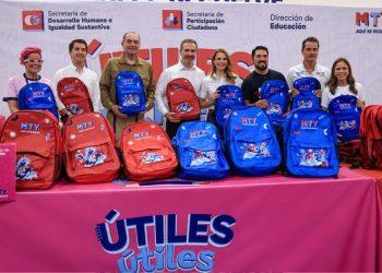 Group of eight people posing with red and blue MTY backpacks for a school supplies drive, standing behind a pink 'ÚTILES' banner.