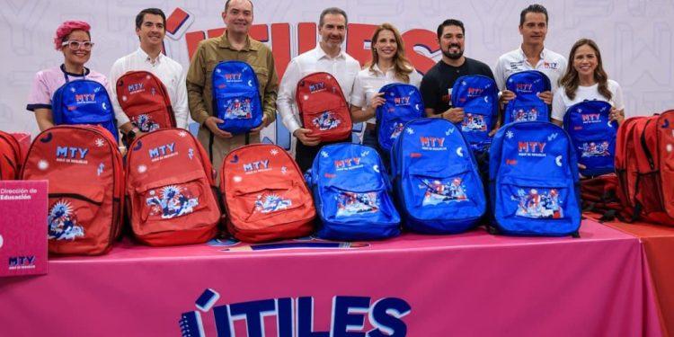 Group of Eight People Posing with Red and Blue Mty Backpacks for a School Supplies Drive, Standing Behind a Pink 'Útiles' Banner. - Apoyo Regio