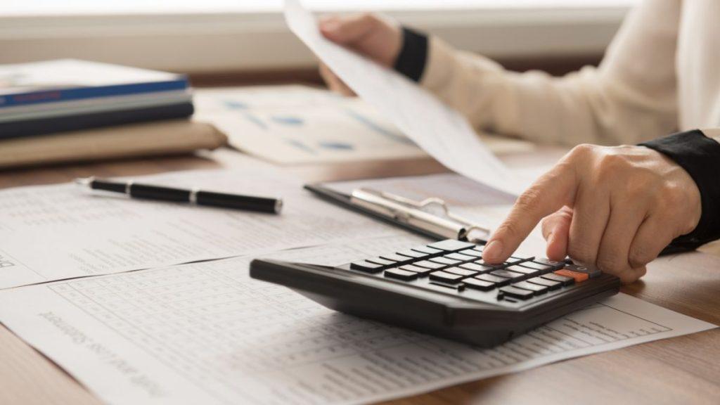 Persona Utilizando Una Calculadora En Un Escritorio Con Documentos Impresos Y Un Bolígrafo Cerca; Probablemente Esté Realizando Tareas De Presupuestación O Contabilidad. - Apoyo Regio