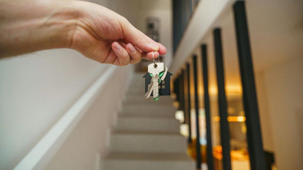 Mano Sosteniendo Unas Llaves En Frente De Un Inmobiliario Donde Vemos Escaleras  - Apoyo Regio