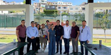Group of Twelve Adults Posing Under a Shaded Pavilion, Standing in a Line with a Green Fence and Apartment Buildings in the Sunny Background. - Apoyo Regio
