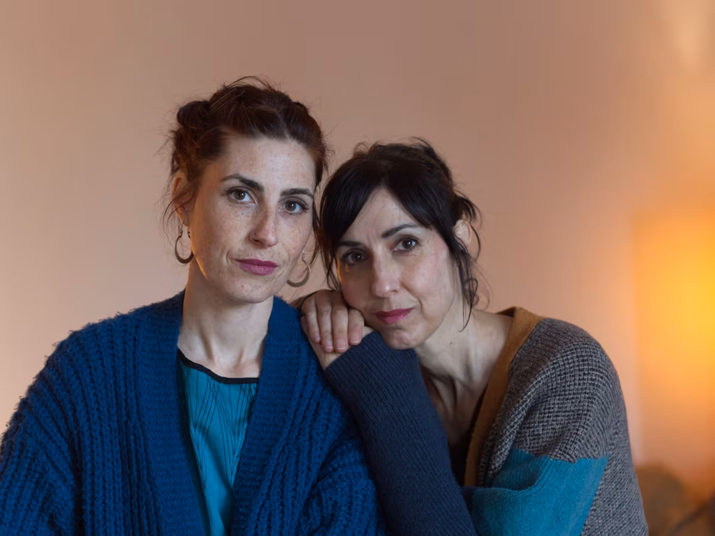 Two Women Sit Close Together, One Resting Her Head on the Other's Shoulder, Both Looking at the Camera with a Calm Expression. - Apoyo Regio