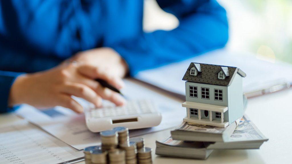 Miniature House on a Stack of Cash with Coins and a Calculator for Mortgage Planning in a Finance Scene - Apoyo Regio