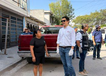 Group of adults standing on a sunny sidewalk beside a red pickup truck; a man in a white shirt with gloves and sunglasses speaks to others nearby.