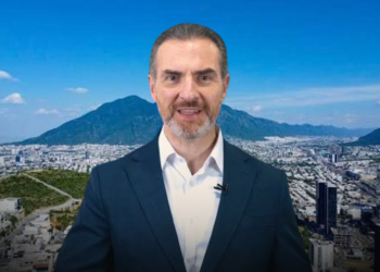 Professional man in a navy suit speaking in front of a city skyline with mountains in the background, wearing a clip-on microphone.