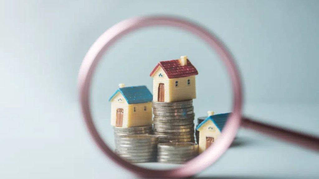 Toy Houses Stacked on Coins, Framed by a Magnifying Glass, Illustrating Real Estate Investment or Housing Costs. - Apoyo Regio